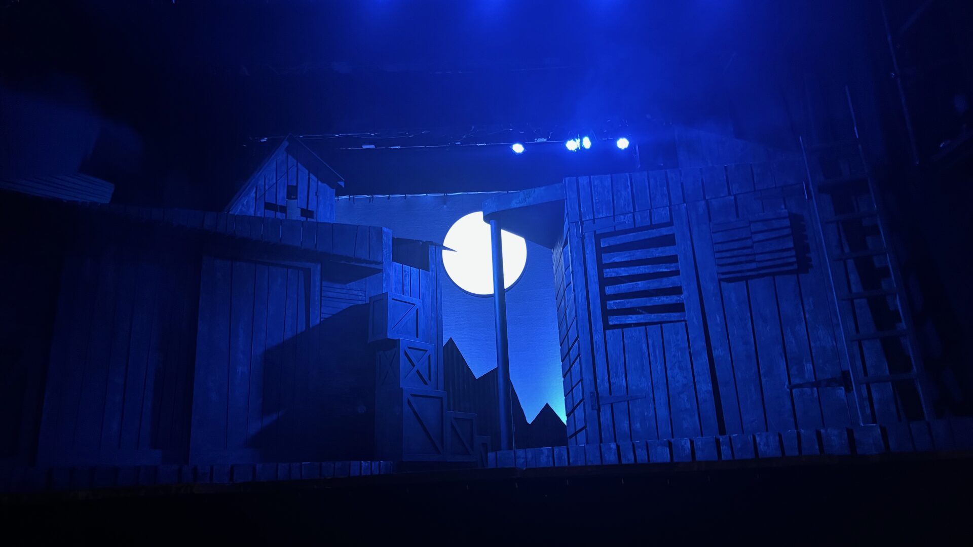 The stage set of Dark of the Moon at the Charing Cross Theatre, bathed in deep blue light. Wooden barn-like structures flank either side of the stage, with a large, luminous full moon glowing at the centre between the buildings. Silhouettes of rooftops and distant mountains are visible in the background, creating an eerie, atmospheric night scene.
