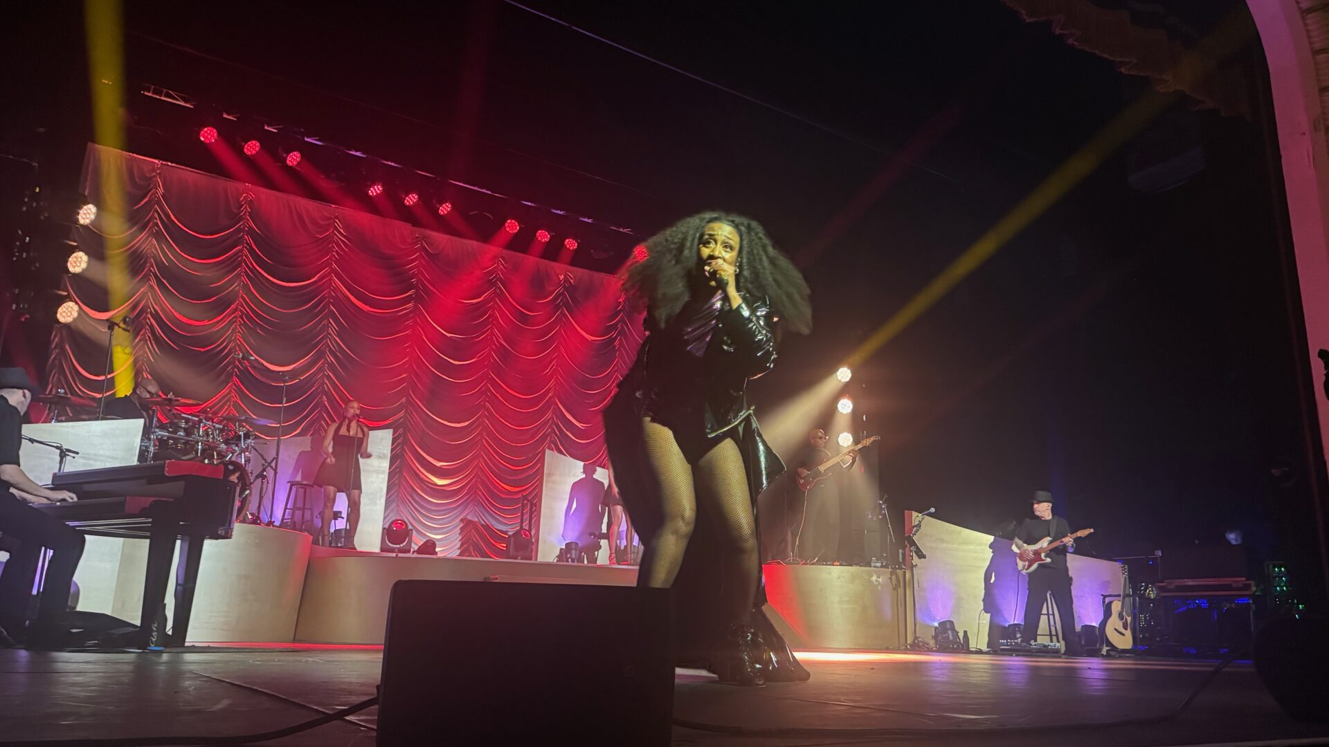 Beverley Knight performing on stage at a concert in Blackpool as part of her Born To Perform Tour. She stands centre stage wearing a black leather outfit with fishnet tights and heeled boots, holding a microphone. Behind her, a full band is visible, including keyboard, guitar, and drum players, beneath dramatic red stage lighting and a swagged curtain backdrop.