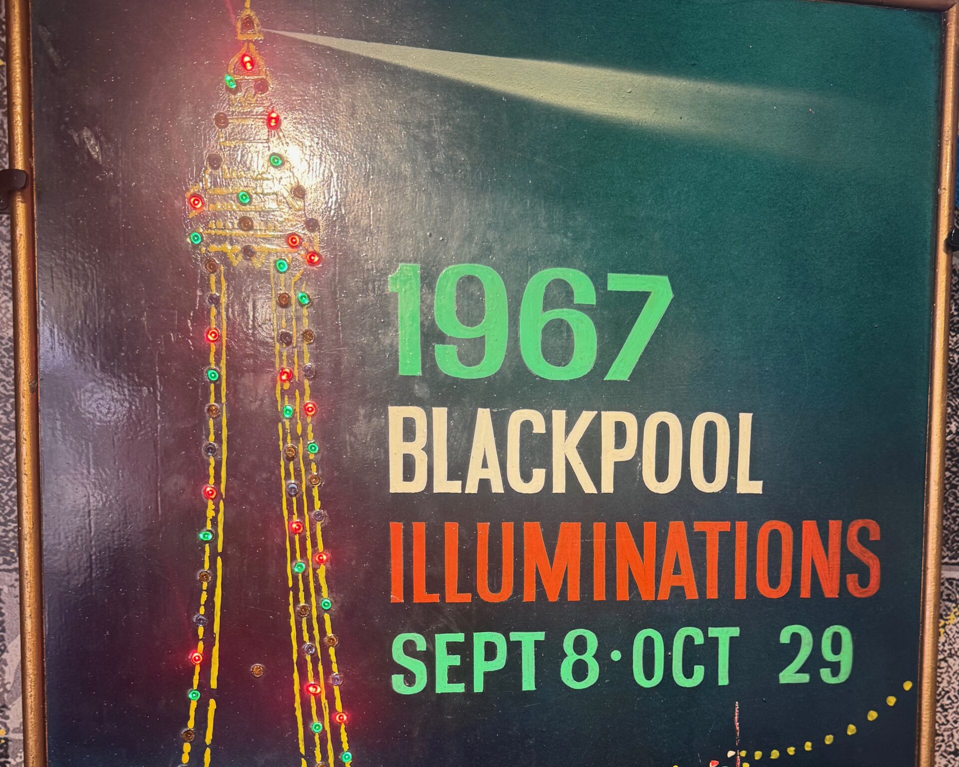 A vintage promotional sign for the 1967 Blackpool Illuminations, running from 8th September to 29th October. The painted board features Blackpool Tower adorned with strings of coloured lights cascading downwards, set against a teal background with a searchlight beam. The text appears in green, white, and orange lettering. The sign is displayed at the Showtown Blackpool museum.