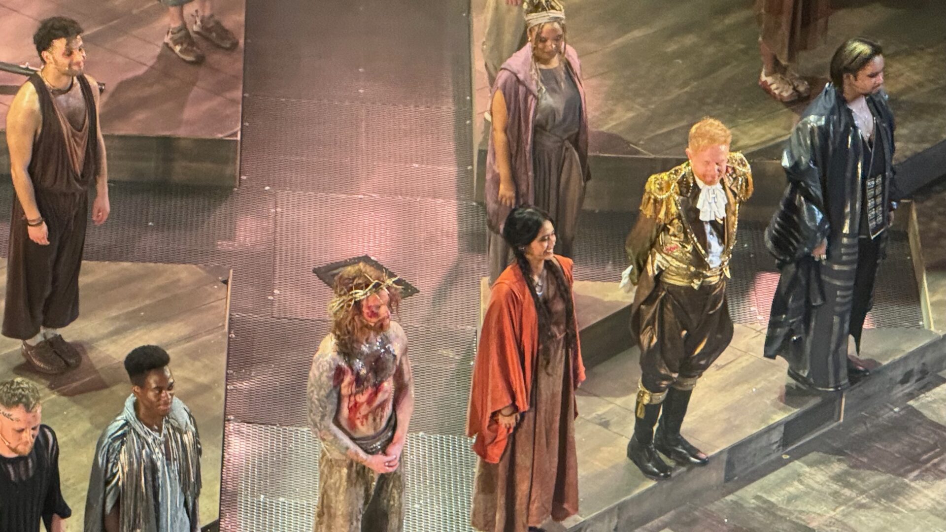 Cast members of Jesus Christ Superstar take their bows on stage at the London Palladium. Photographed from above, the ensemble includes Sam Ryder in the role of Jesus wearing a crown of thorns and a blood-streaked costume, Jesse Tyler Ferguson in an ornate gold military-style jacket representing Herod, a woman in an orange robe, and several other cast members in period and contemporary costumes, all lined up across the stage.