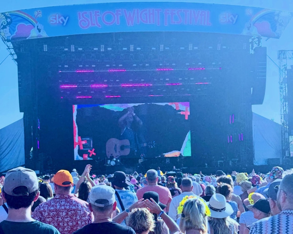 A large crowd of festival-goers watches Amy McDonald perform on the main stage at the 2025 Isle of Wight Festival. The stage features vibrant pink and blue lighting effects with "ISLE OF WIGHT FESTIVAL" prominently displayed across the top, along with Sky branding. The performer can be seen on stage with an acoustic guitar, backed by colourful visual displays. The audience, wearing a mix of caps, hats, and summer clothing, fills the foreground of the image, creating the typical atmosphere of a major music festival performance.