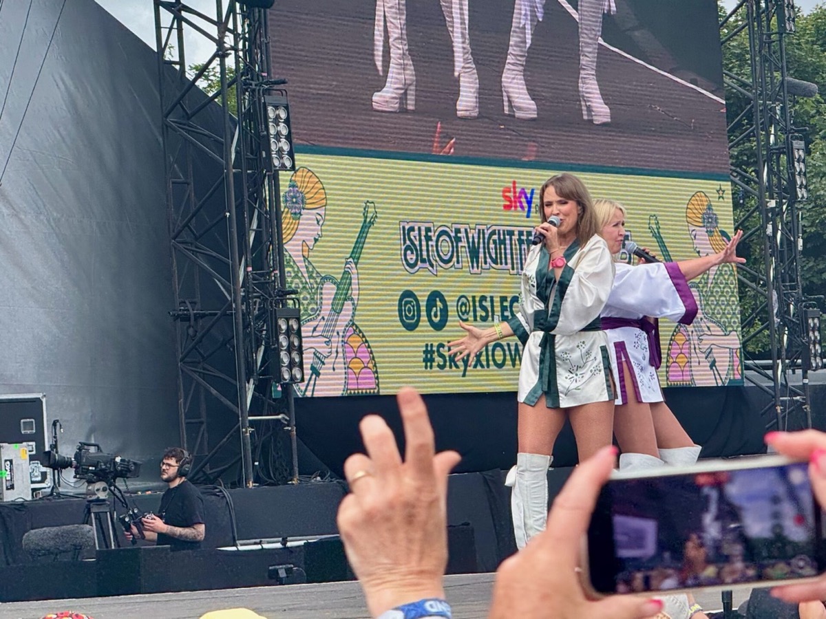 Bjorn Again performs on stage at the 2025 Isle of Wight Festival, with two female performers in white outfits singing into microphones. The stage backdrop features colourful retro-style graphics and the Isle of Wight Festival branding with Sky sponsorship visible. In the foreground, audience members can be seen with their hands raised and mobile phones recording the performance. A photographer is positioned in the pit area capturing the show, whilst stage rigging and lighting equipment frame the scene. The vibrant stage design creates an energetic atmosphere typical of the ABBA tribute act's performances.