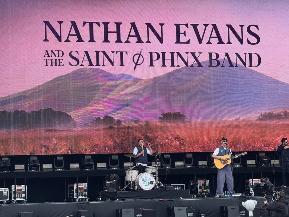 Nathan Evans and the Saint PHNX Band perform on a large outdoor festival stage. A drummer and vocalist stand at microphones to the left, whilst Nathan Evans stands to the right playing an acoustic guitar and singing. Behind them, a large LED screen displays the text "Nathan Evans and the Saint PHNX Band" above a dramatic landscape of purple hills and golden meadows. The drum kit bears the band's logo.