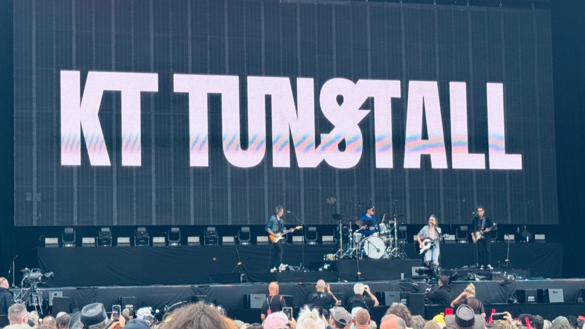 KT Tunstall and her band perform on a large outdoor festival stage. KT, wearing a denim jacket and holding an acoustic guitar, stands at a microphone to the right of centre. A guitarist stands to her left, with a drummer in a blue top seated behind a white kit at the centre rear, and a second guitarist on the far right. A vast LED screen behind them displays "KT Tunstall" in bold lettering. The crowd in the foreground holds up phones to capture the moment.