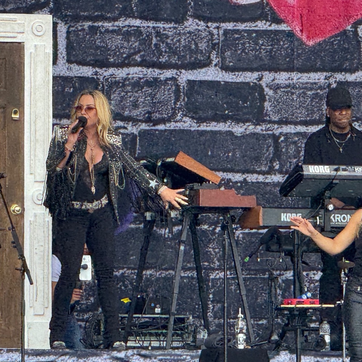 Singer Anastasia performs on an outdoor festival stage, dressed in a black sequinned fringed jacket, black trousers and rose-tinted sunglasses, with layered necklaces and long blonde hair. She holds a microphone in one hand. A keyboard player wearing a black cap stands at a Korg Kronos synthesiser to her right. The backdrop features a graffiti-style brick wall design.