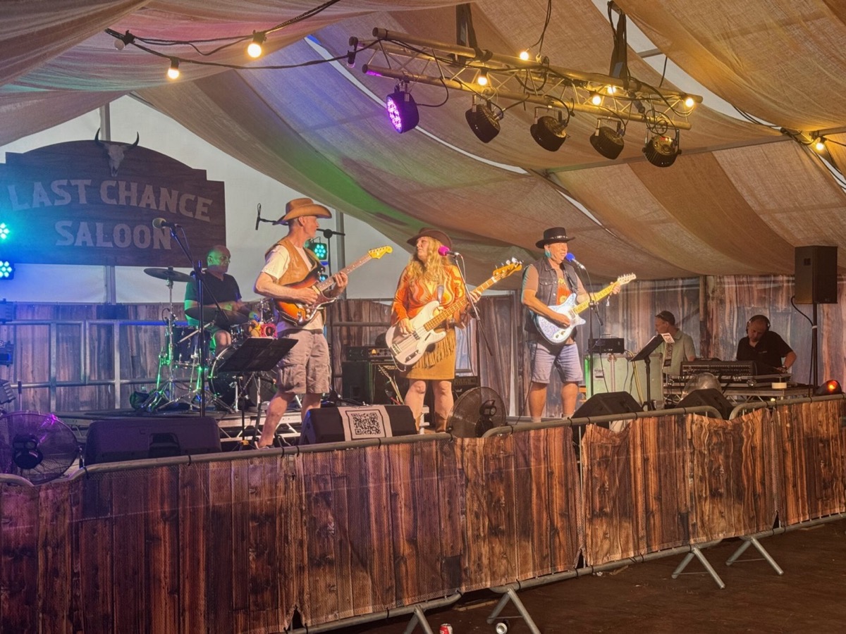 A four-piece band performs inside a marquee tent decorated as the Last Chance Saloon, with a rustic wooden sign bearing that name visible on the left. Three guitarists and a female vocalist in an orange-patterned dress and a wide-brimmed hat are spread across the stage, along with a drummer visible at the rear. A sound engineer is visible to the right. String lights and colourful stage lighting illuminate the canvas ceiling above.