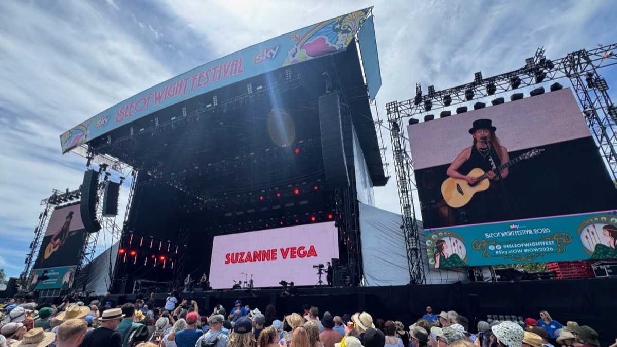 A wide shot of the Sky-sponsored Isle of Wight Festival main stage during Suzanne Vega's performance on a sunny afternoon. A central LED screen displays her name in bold red lettering. A side screen shows a close-up of Suzanne performing, wearing a black top hat, black sleeveless top, and gold layered necklaces whilst playing an acoustic guitar. The large crowd in the foreground wear hats and summer clothing, many holding up phones to capture the moment. The festival's colourful branded canopy is visible above the stage.