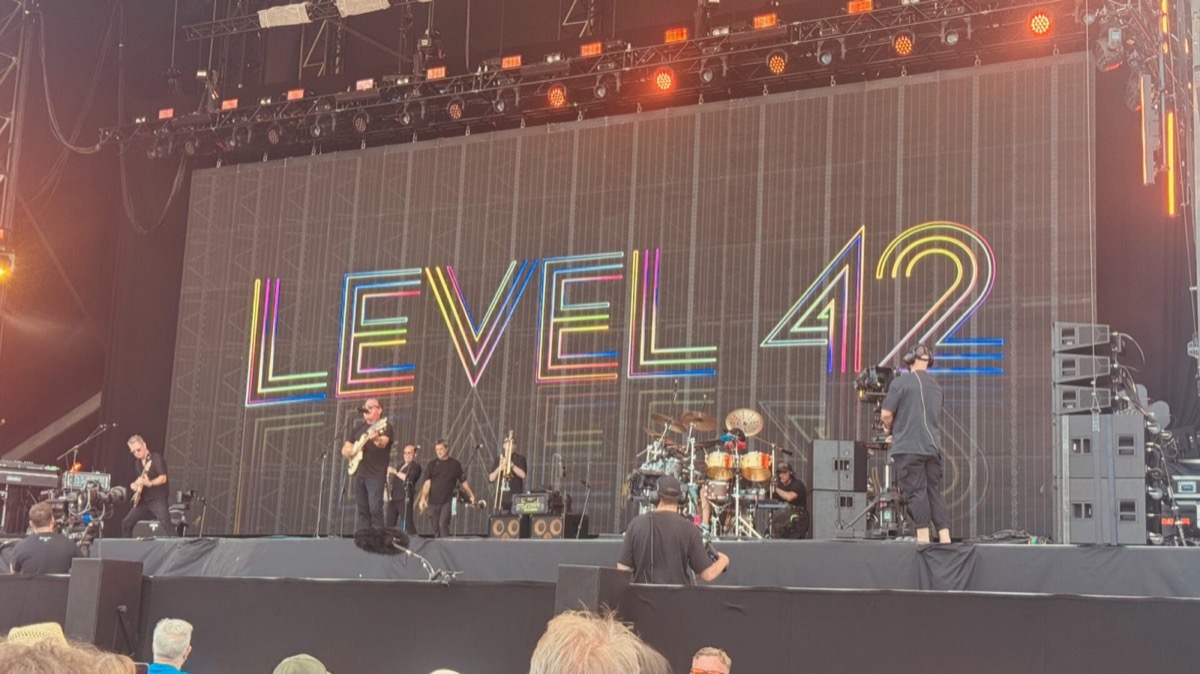 Level 42 perform on a large outdoor festival stage, with the band's name displayed on the LED backdrop in multicoloured neon-style lettering outlined in rainbow hues. Several band members dressed in black are spread across the stage — a bassist prominent at the front, with horn players, a drummer behind a large kit with gold cymbals, and other musicians visible. Camera operators are positioned at the front and side of the stage. A warm orange light glows from stage left.
