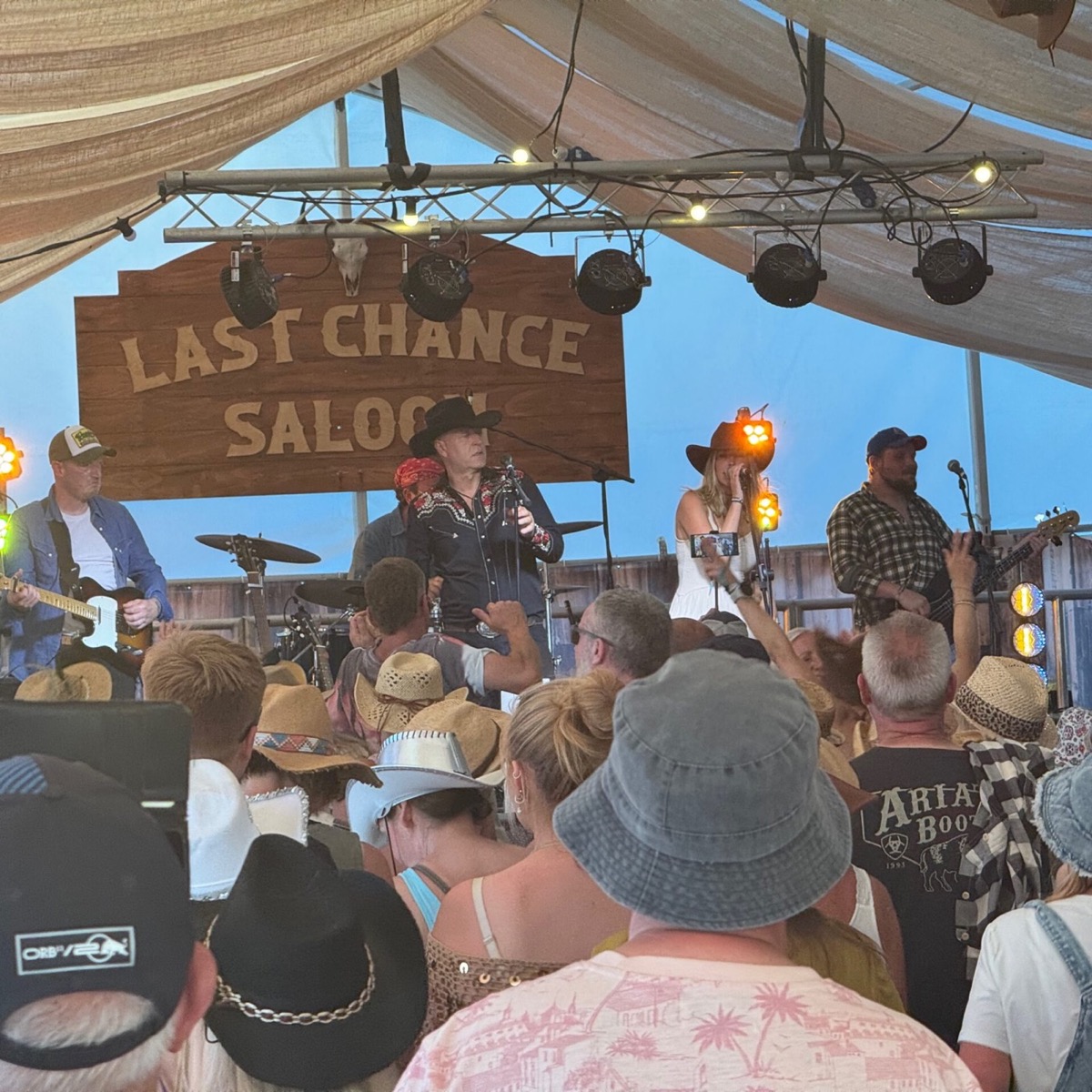 A country band performs on the Last Chance Saloon stage inside a marquee tent at the Isle of Wight Festival. A large wooden sign reading "Last Chance Saloon" with a mounted skull hangs prominently above the stage. The band includes a male vocalist in a black cowboy hat and embroidered western shirt, a female vocalist in a white dress and cowboy hat, a guitarist in a checked shirt, and a further guitarist on the left. The audience in the foreground is densely packed, with many wearing cowboy and wide-brimmed hats. Warm amber stage lights illuminate the scene against an early evening sky visible through the open sides of the tent.