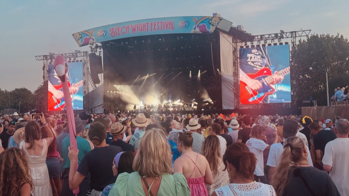 A large crowd faces the Isle of Wight Festival main stage at dusk during The Cure's headline performance. The stage is filled with dramatic white haze and beams of light. The two flanking LED screens display close-ups of a red electric guitar being played, alongside text referencing the band. Trees are visible on the horizon against a pale evening sky. A pink inflatable flamingo can be seen rising above the crowd on the left.